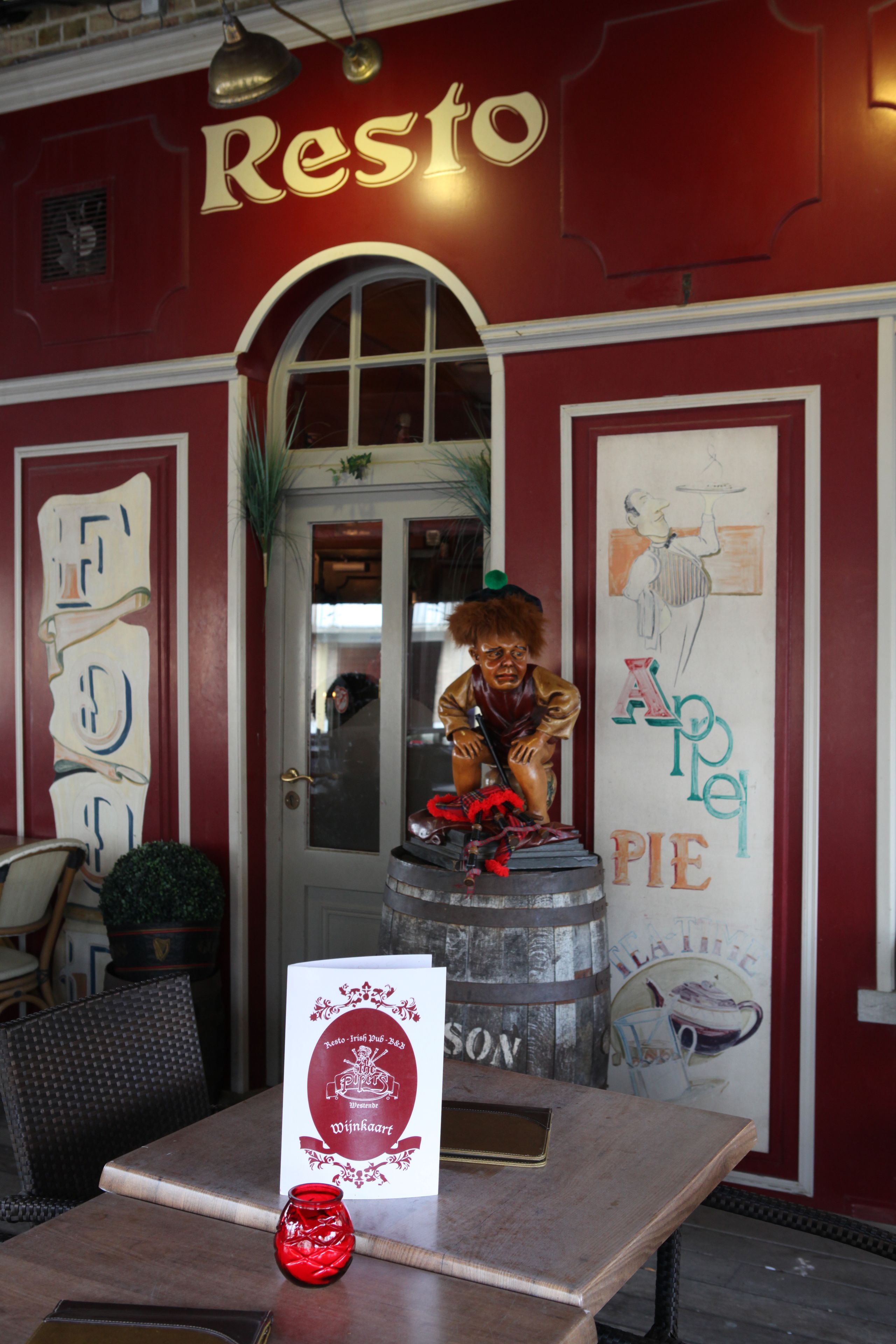 Ein Restaurant Eingang mit roter Fassade, einem großen Bogenfenster und einer Statue einer Person auf einem Fass.