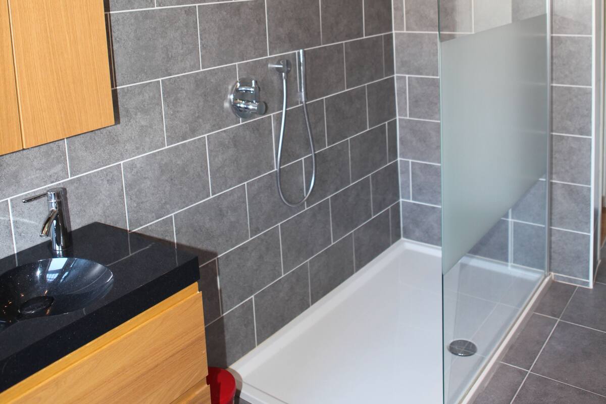 A modern bathroom with a glass-enclosed shower, a wooden vanity, and grey tiled walls.