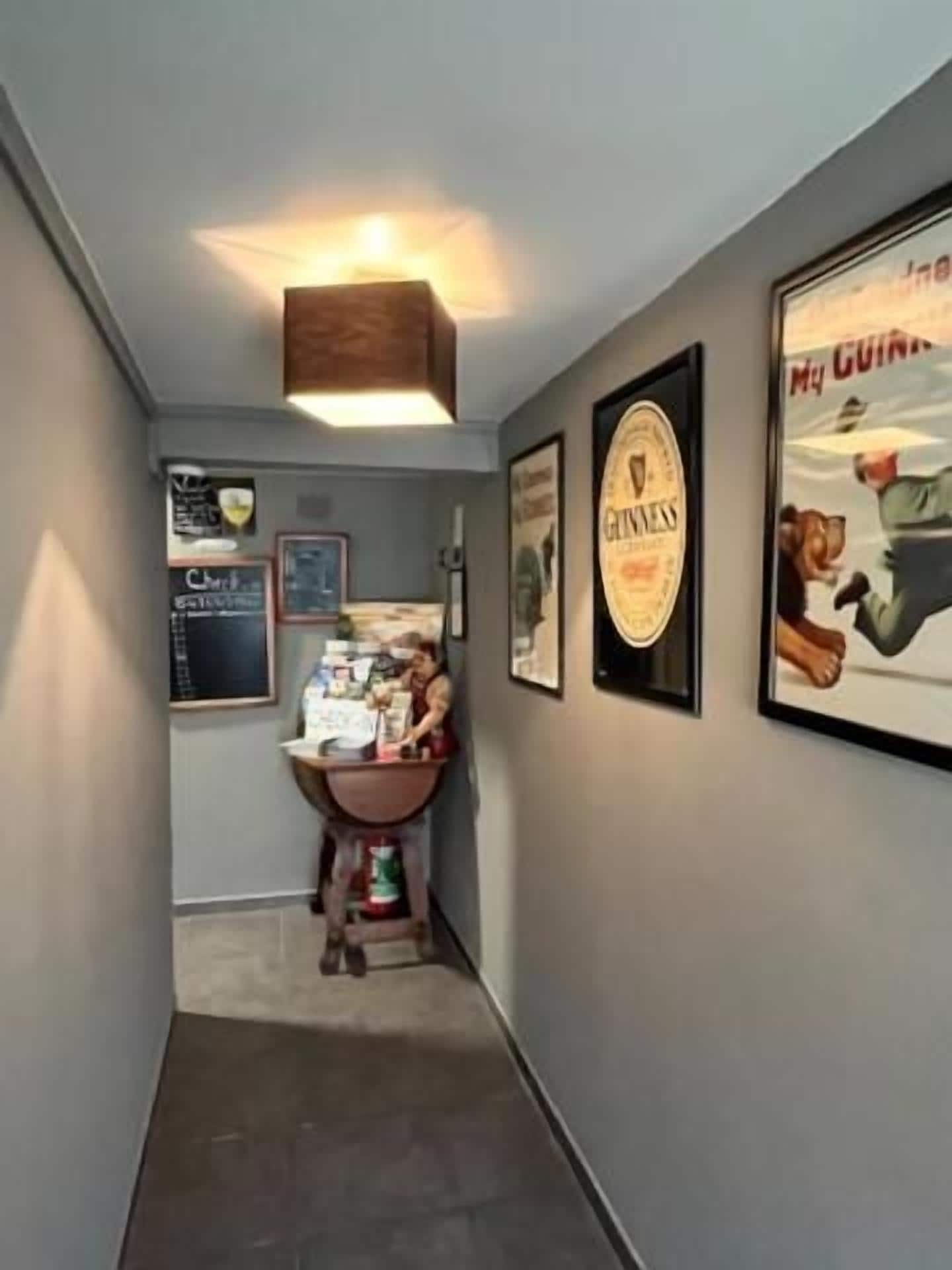 A narrow hallway with a wooden ceiling light fixture, a small table with various items, and framed posters on the walls.