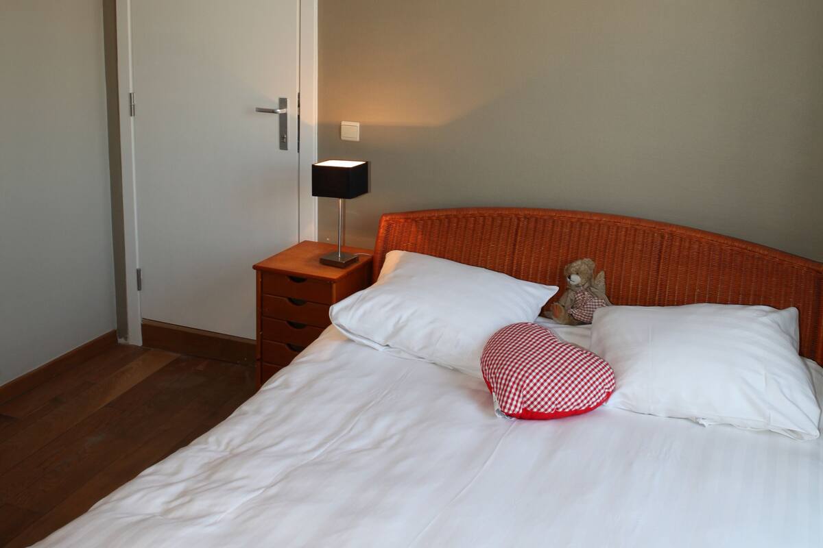 A neatly made bed with white linens, a red and white checkered pillow, and a small teddy bear beside it. There is a bedside table with a lamp and a door in the background.