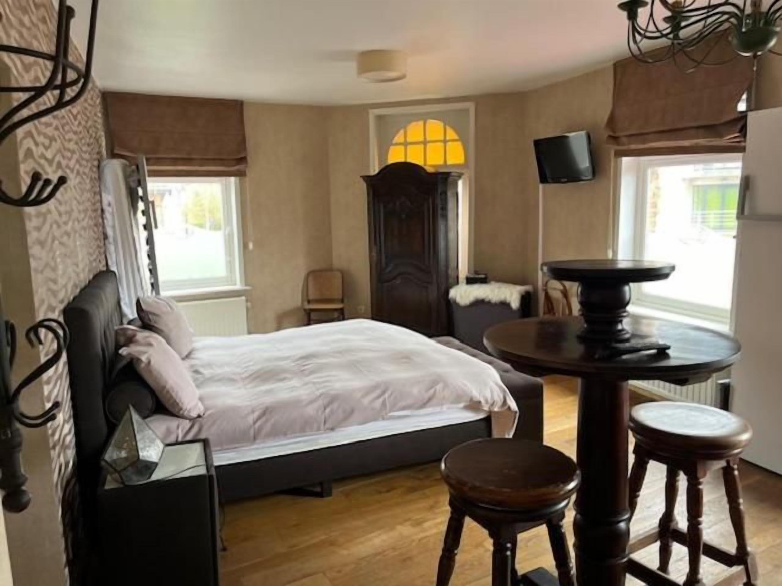 Ein Schlafzimmer mit einem Bett, einem runden Tisch mit Hocker, einem Fernseher und einem Fenster mit Vorhängen.