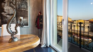 Una habitación con una mesa de madera, una escultura decorativa de metal, un maniquí con una chaqueta roja, y vista a un canal con barcos a través de una ventana con barandilla de hierro forjado.
