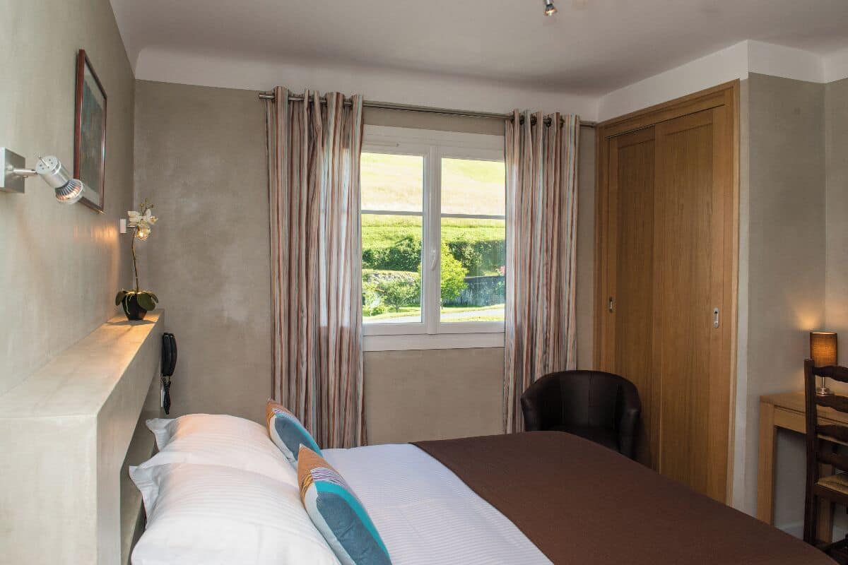 A bedroom with a bed, a window with curtains, a bedside lamp, a chair, and a wooden wardrobe.