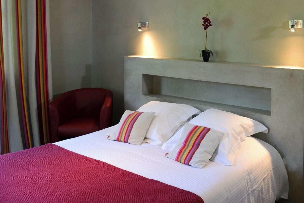 A hotel room with a bed, a red chair, a wall-mounted shelf with a potted plant, and two wall sconces.