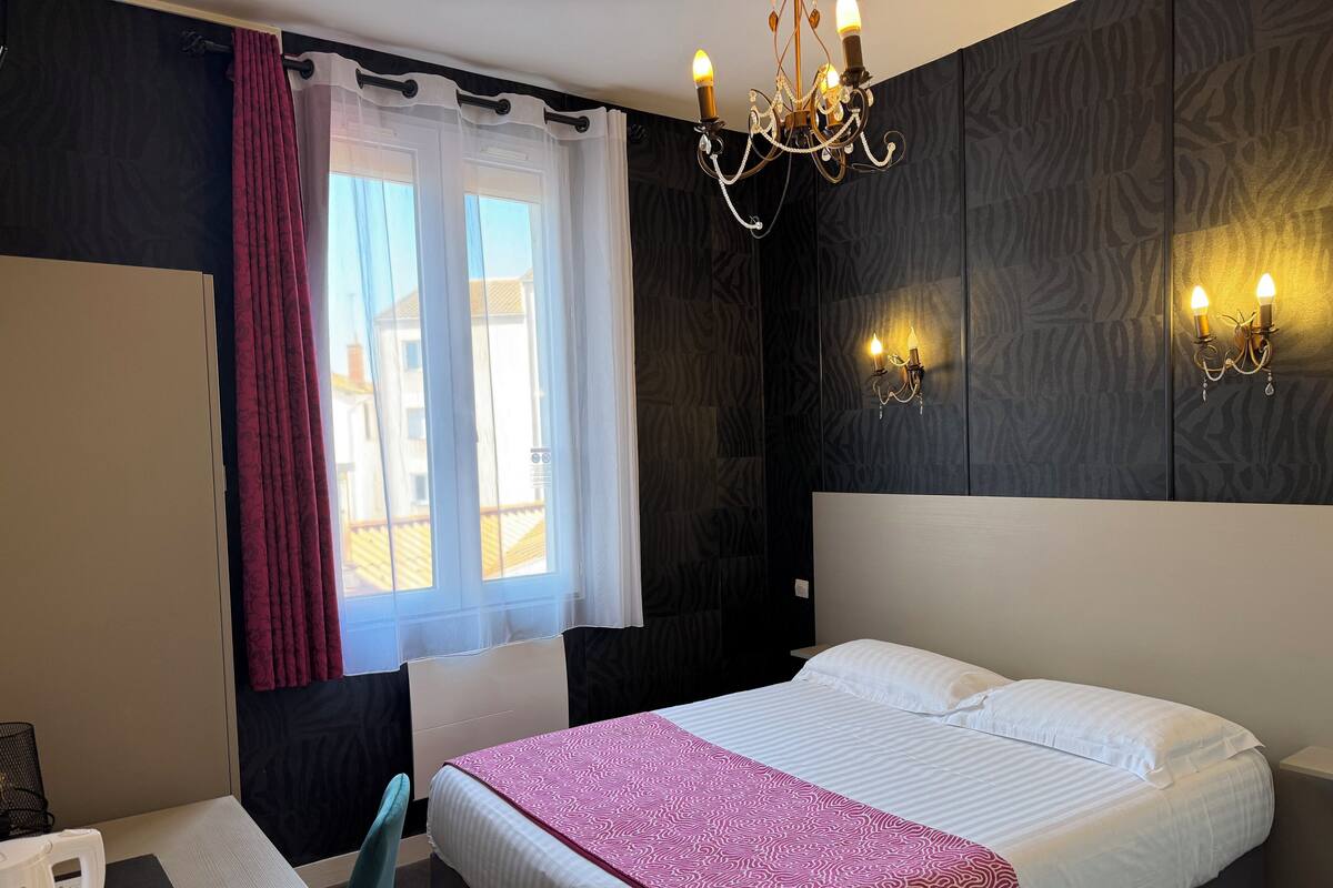 Hotellihuone, jossa on sänky, työpöytä, tuoli, ikkuna verhoilla ja seinään kiinnitetyt valaisimet.