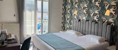 A hotel room with a bed, a desk, a chair, a radiator, and a view of the outside balcony.