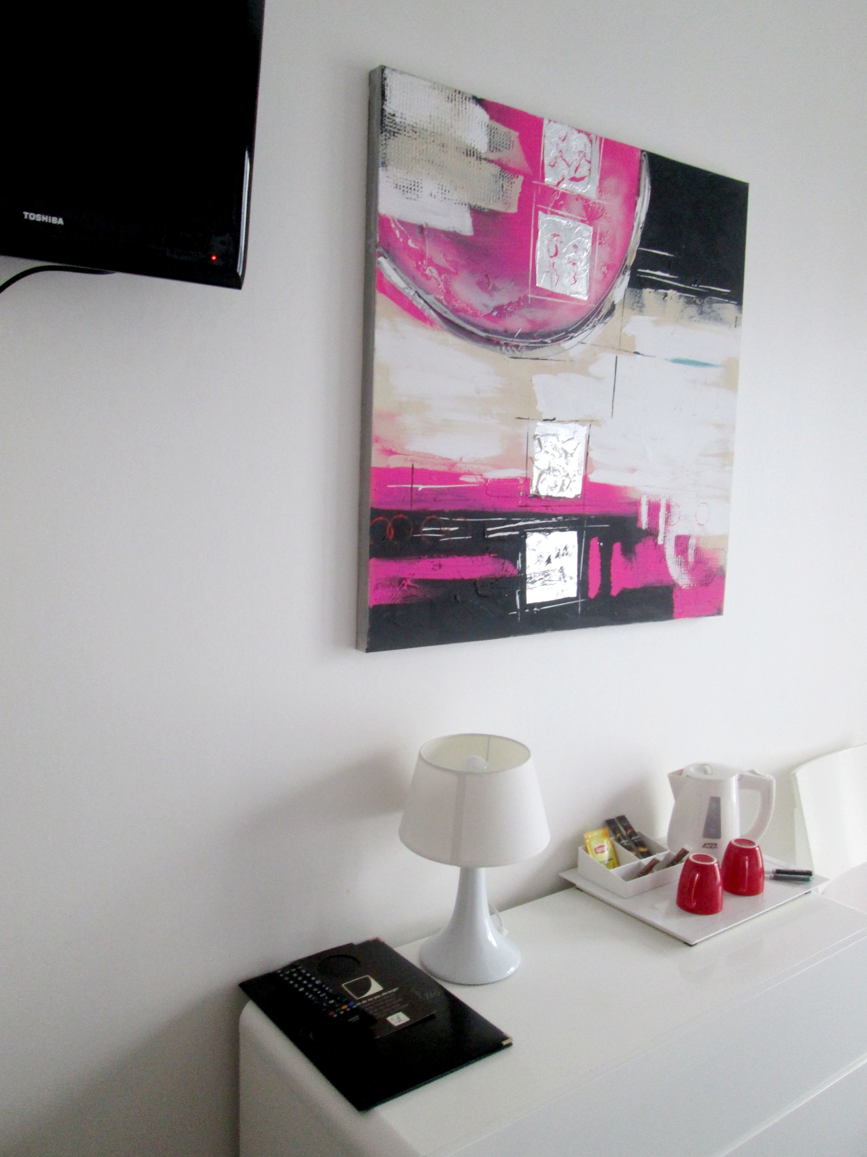 A hotel room with a white wall, a flat-screen TV mounted above a white table, a painting with abstract pink and black colors, a white lamp, and a tray with red cups and snacks.