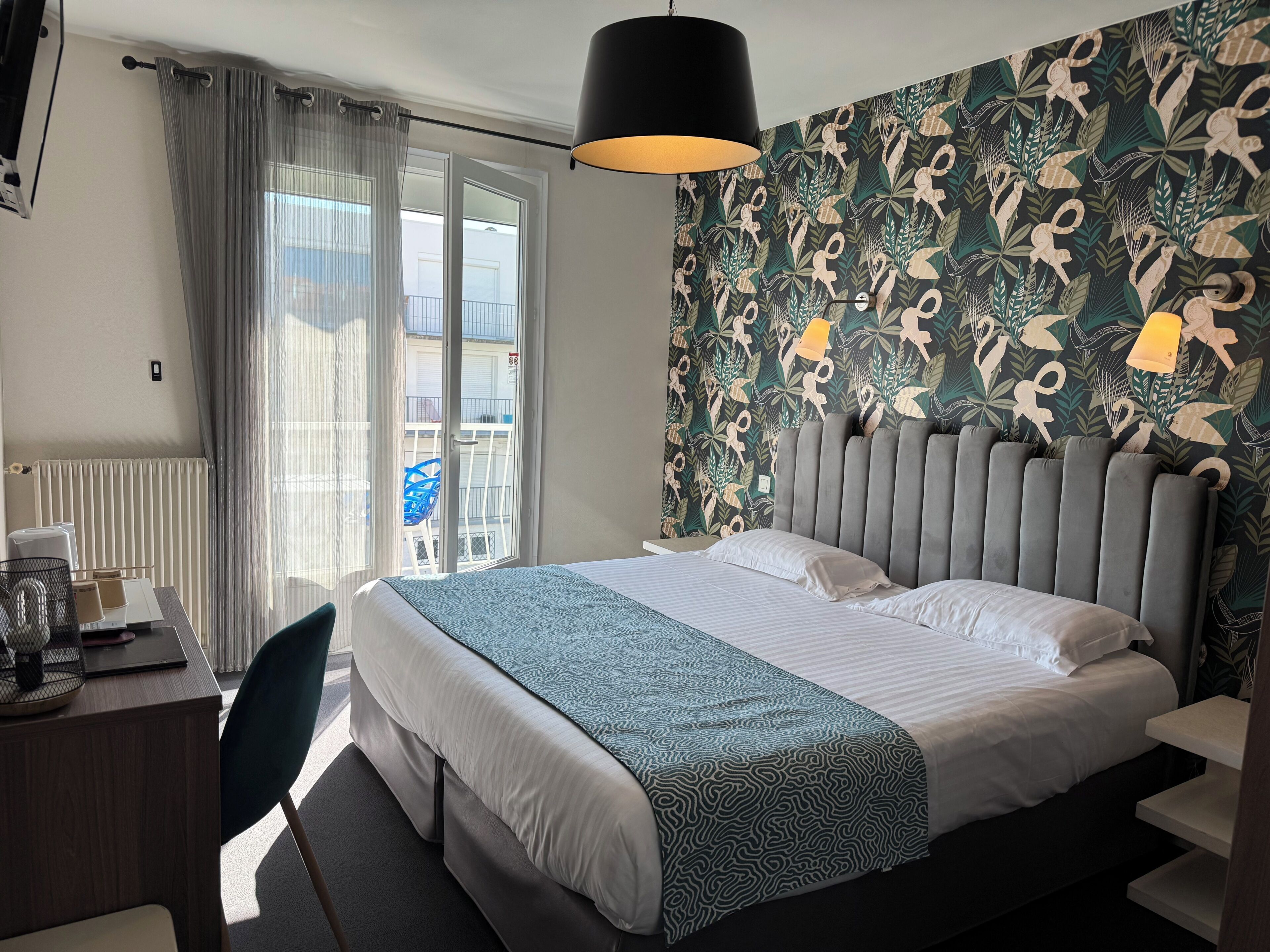 A hotel room with a bed, a desk, a chair, a radiator, a window with curtains, and a patterned wall.