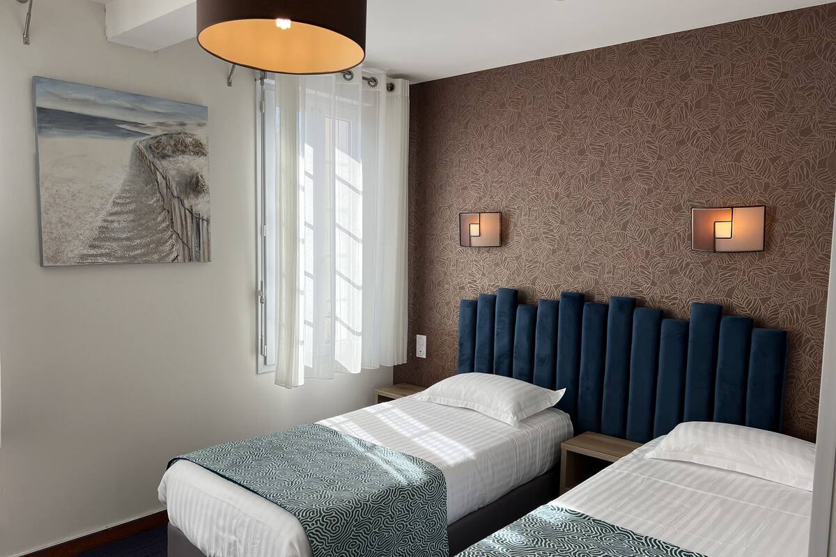 Hotellihuone, jossa on kaksi sänkyä, suuri ikkuna läpikuultavilla verhoilla, seinään kiinnitetty valaisin ja taulu seinällä.
