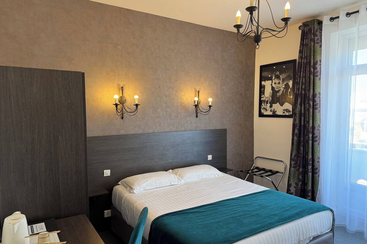 Hotellihuone, jossa on sänky, yöpöydät, työpöytä tuolilla, kattokruunu ja ikkuna, jossa on verhot.