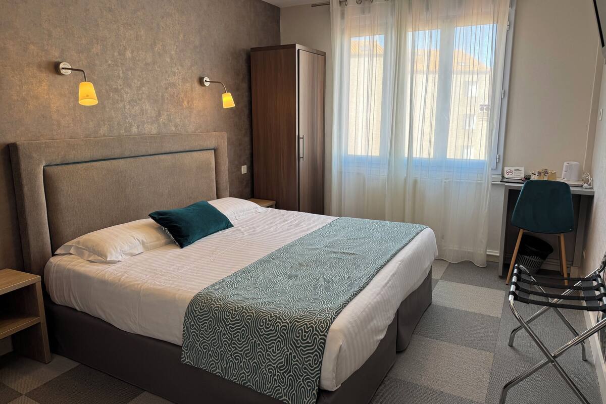 Hotellihuone, jossa on suuri sänky, kaksi yöpöytälamppua, vaatekaappi, työpöytä tuolilla ja näkymä ulos ohuiden verhojen läpi.