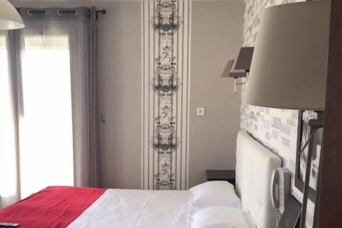 Hotellihuone, jossa on sänky, yöpöytä, jossa on punaisia mukeja, lamppu ja ikkuna, jossa on verhot.
