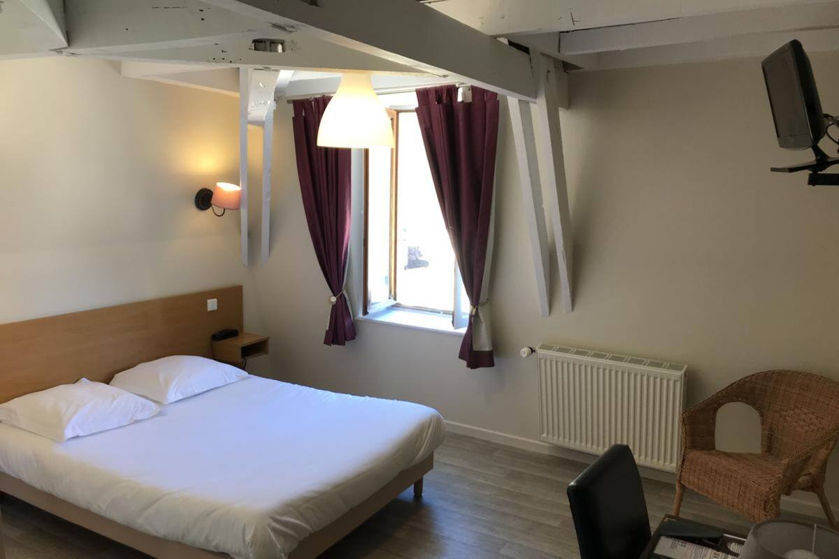 A hotel room with a bed, a chair, a desk with a computer, a window with curtains, and a radiator.