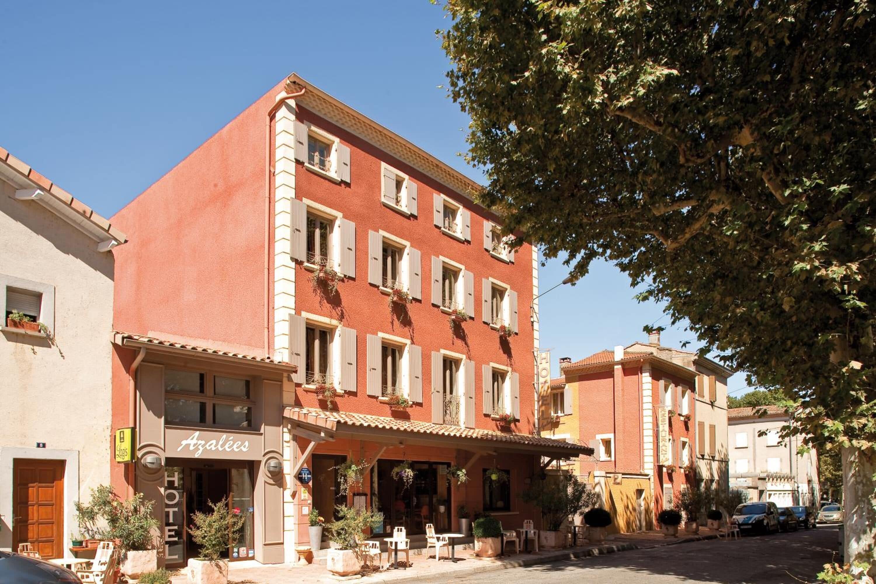 A hotel with a red facade, white shutters, and outdoor seating.