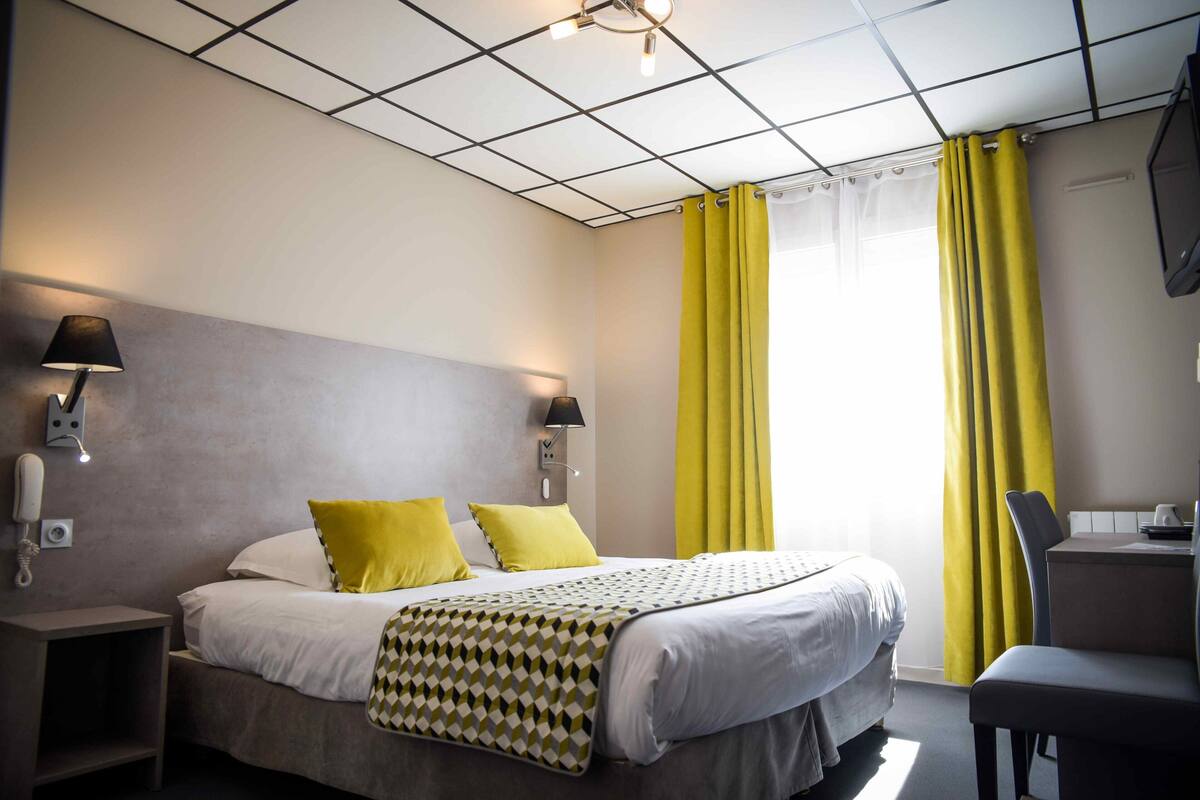 A hotel room with a bed, two bedside lamps, a telephone, a desk with a chair, and a window with yellow curtains.