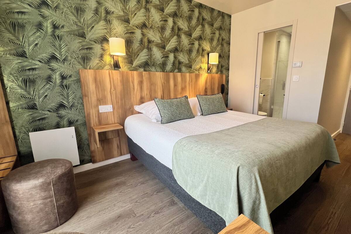 A hotel room with a bed, a nightstand, a chair, a wooden headboard, and a wall with a leaf pattern.