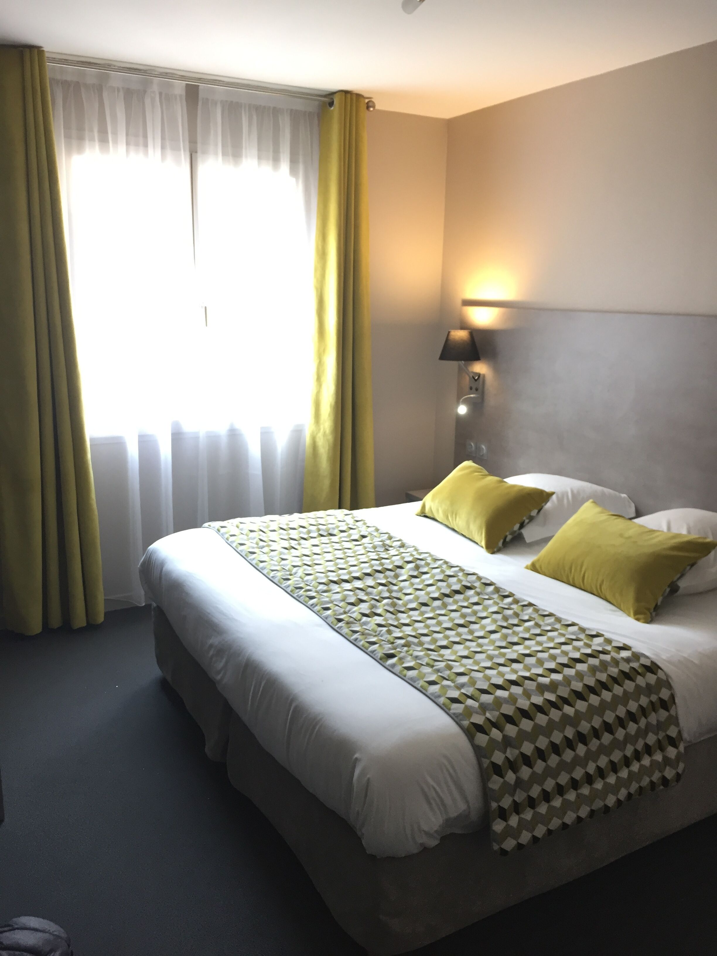 A hotel room with a large bed, yellow pillows, a patterned throw, and a lamp on the wall.
