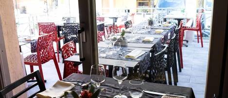 An outdoor dining area with tables set for two, red and black chairs, and a view of the street and buildings in the background.