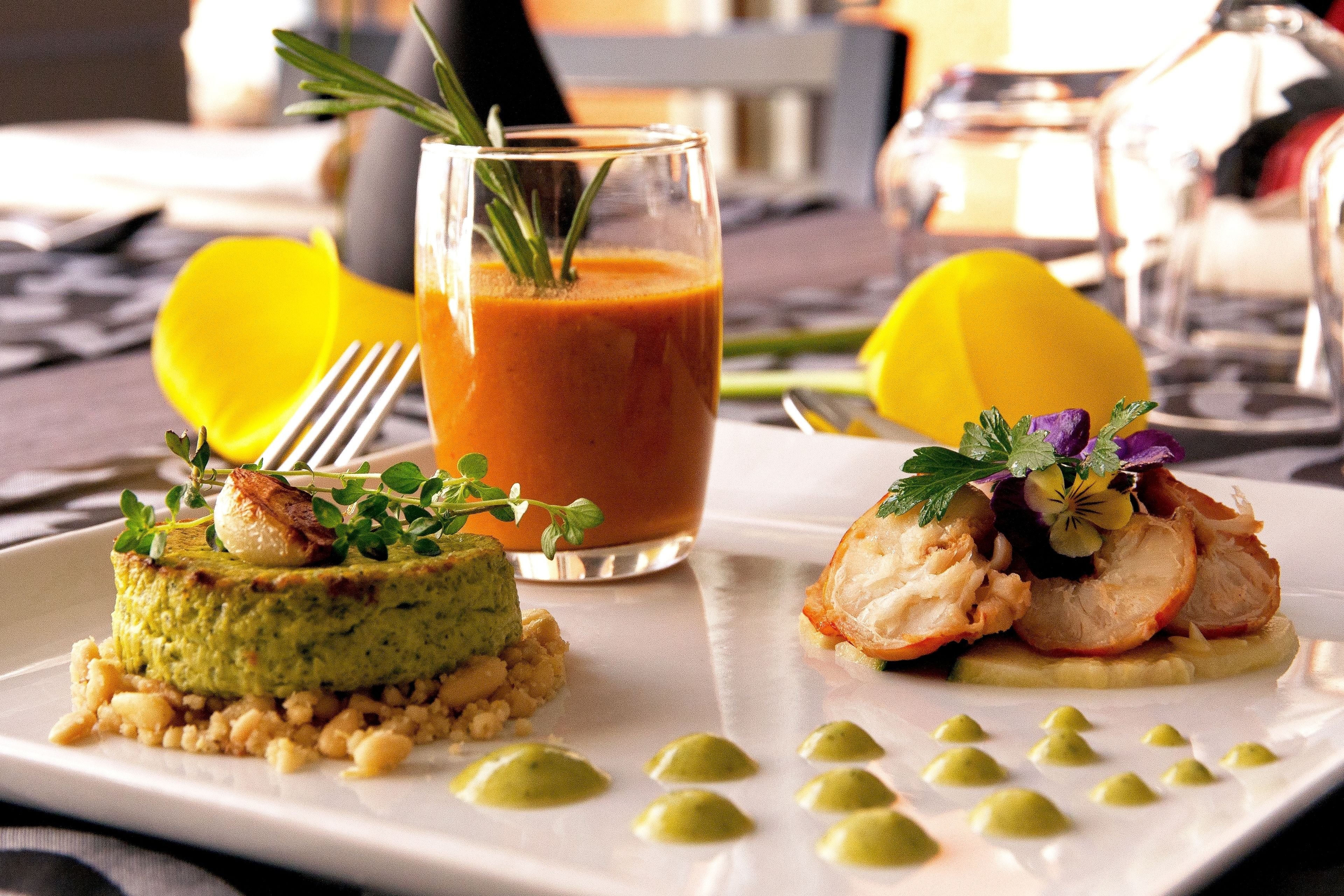 Un plato con un mousse verde, un vaso de líquido rojo con un adorno, y una fruta amarilla sobre un plato blanco.