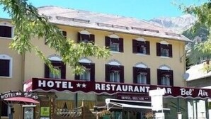 A two-story building with a sign indicating it is a hotel and restaurant. There are outdoor seating areas with umbrellas, and the building has a traditional architectural style with arched windows. - Bel'Air (Jausiers)