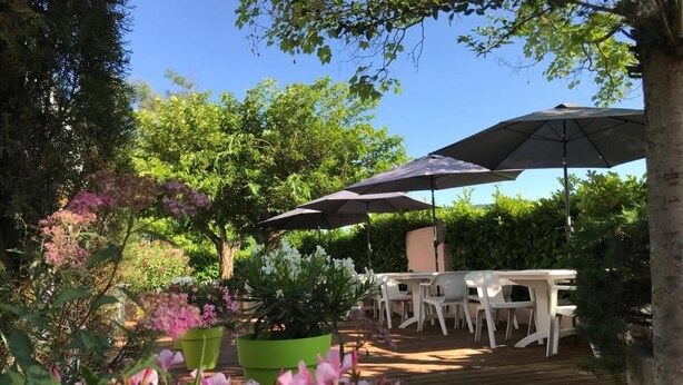 An outdoor patio area with a wooden deck, white plastic tables and chairs, and a large umbrella providing shade.