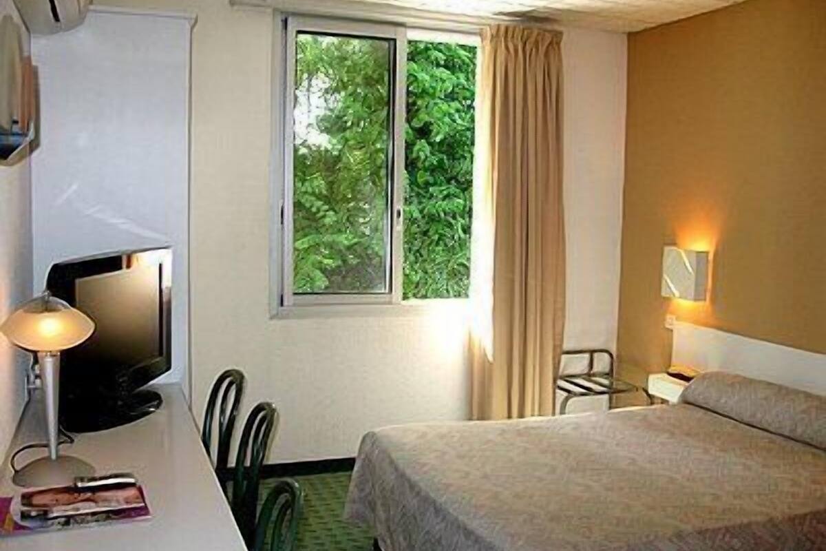 Habitación de hotel con cama, escritorio con televisor, una silla y ventana con cortinas.