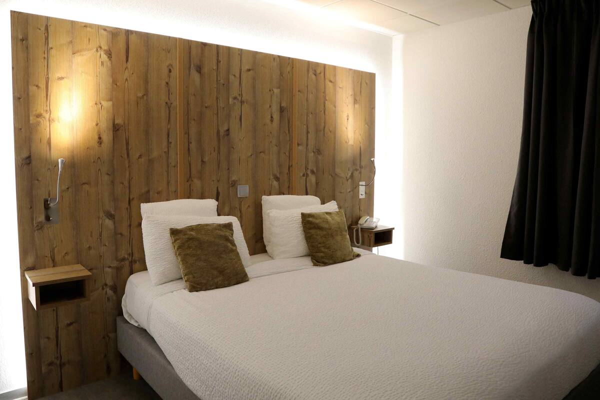 A hotel room with a wooden headboard, a bed with white bedding and two pillows, a bedside table with a lamp, and a telephone.