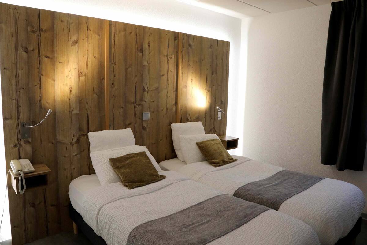 A double bed with white bedding and two brown pillows, a wooden headboard, a bedside table with a lamp, and a telephone.