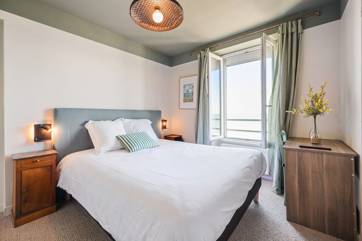 A neatly made bed with white linens and a striped pillow, a bedside table with a lamp, a wooden nightstand, a window with curtains, and a vase with yellow flowers on a wooden dresser.