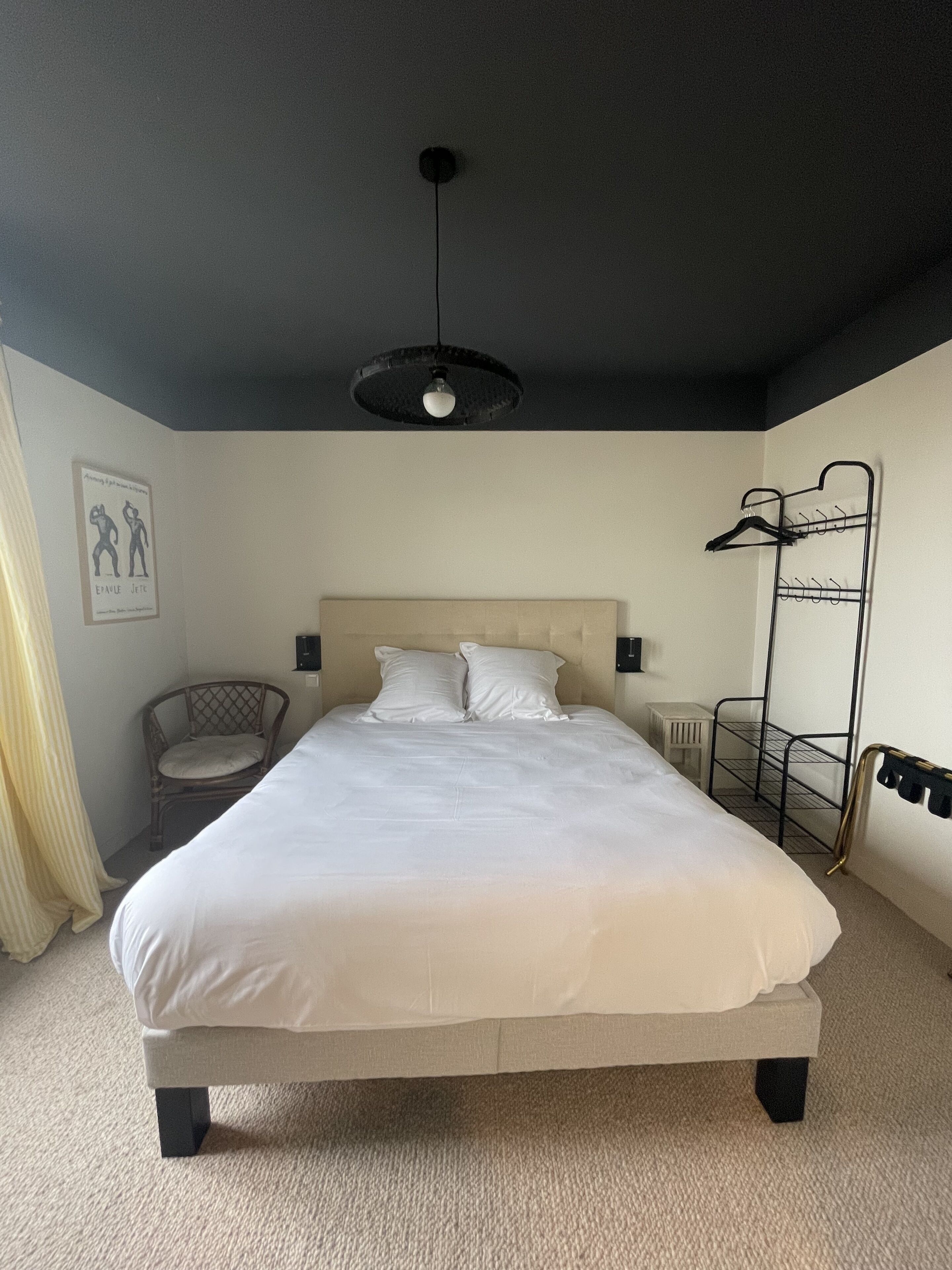 A neatly made bed in a room with a headboard, a chair, a coat rack, and a wall-mounted shelf.