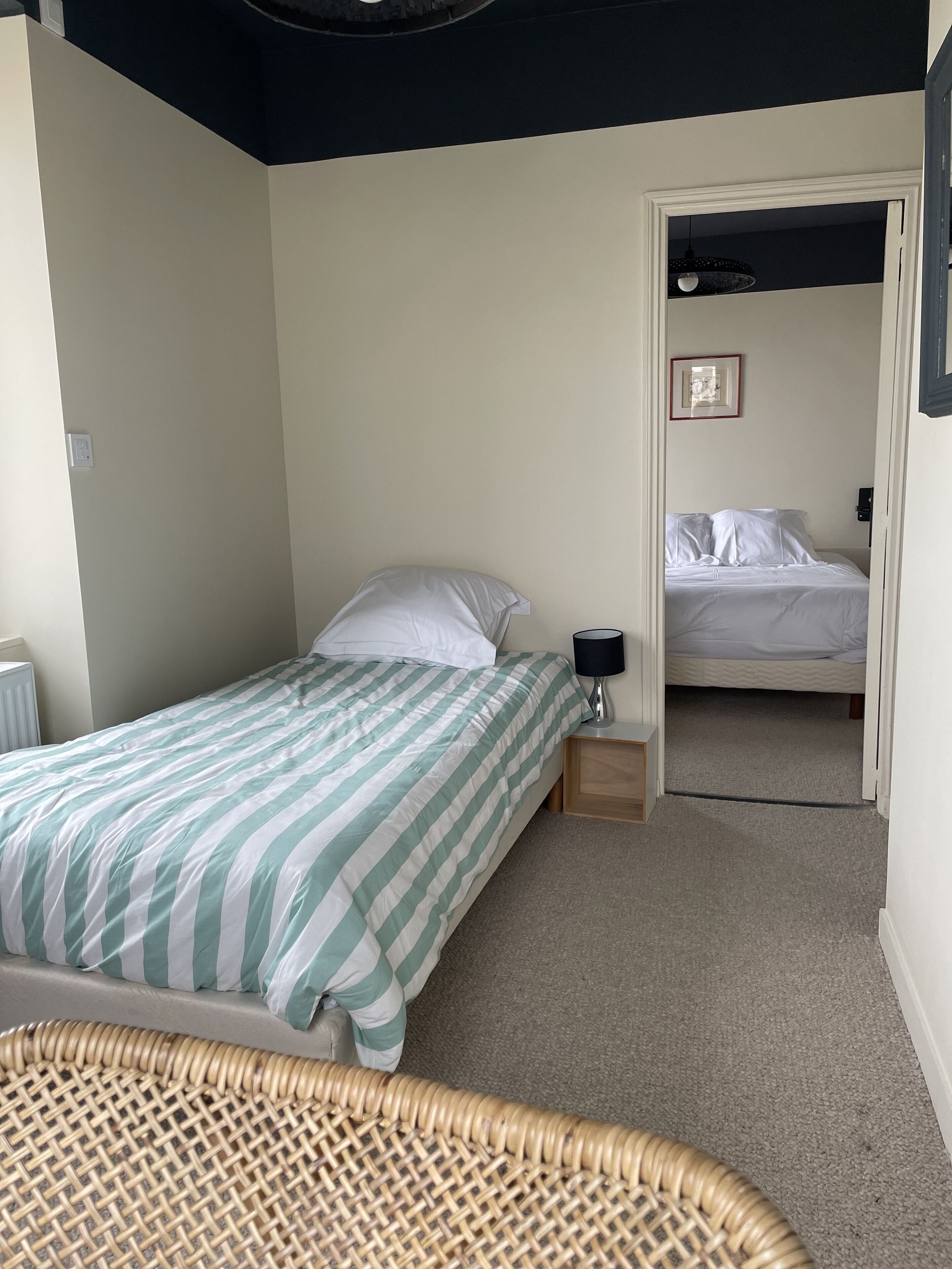 A bedroom with a single bed, a bedside table with a lamp, a small wooden nightstand, and a wicker chair.
