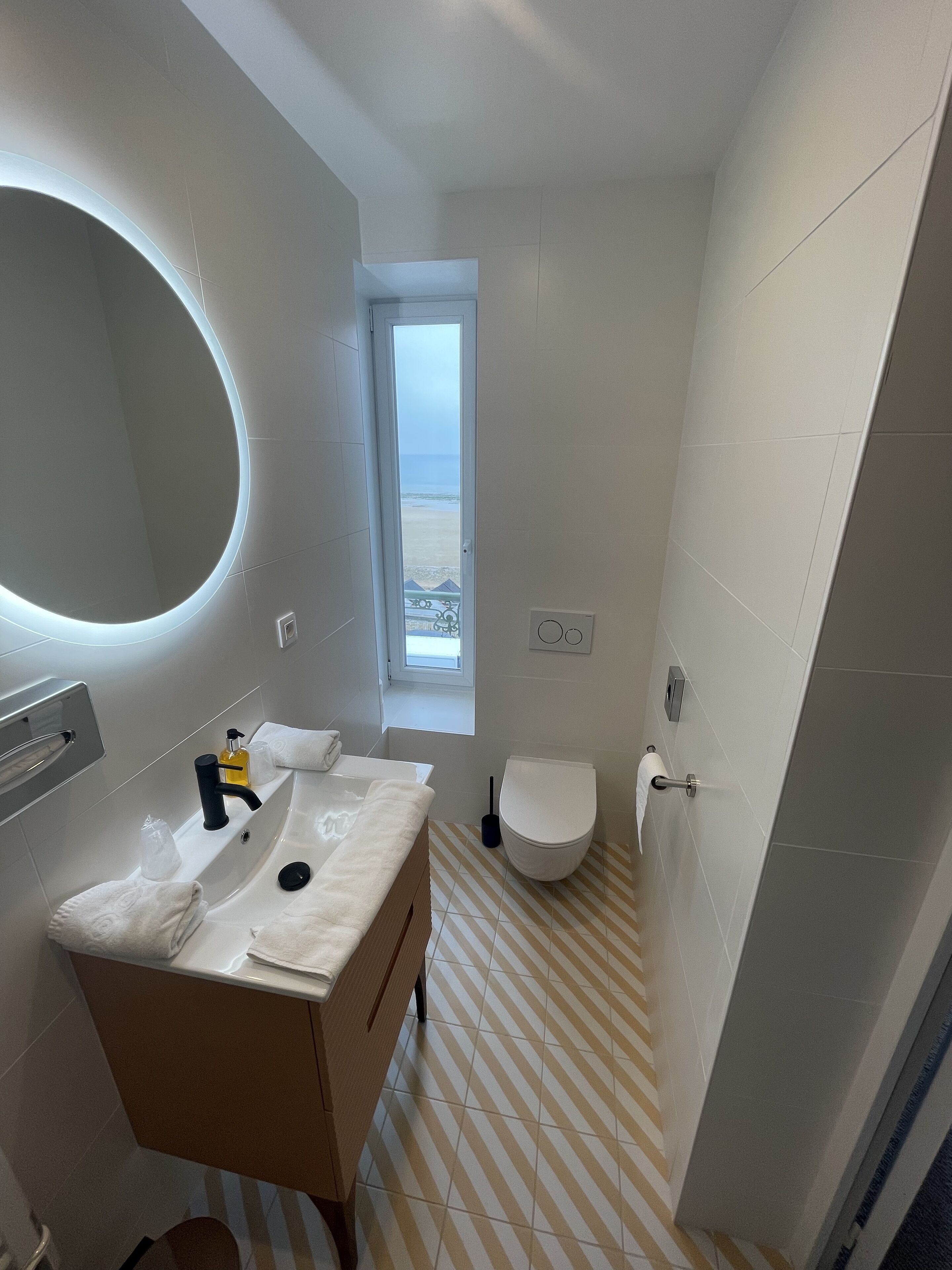 Une salle de bain moderne avec un grand miroir, un lavabo équipé de produits de toilette, des toilettes et une fenêtre donnant sur la plage.