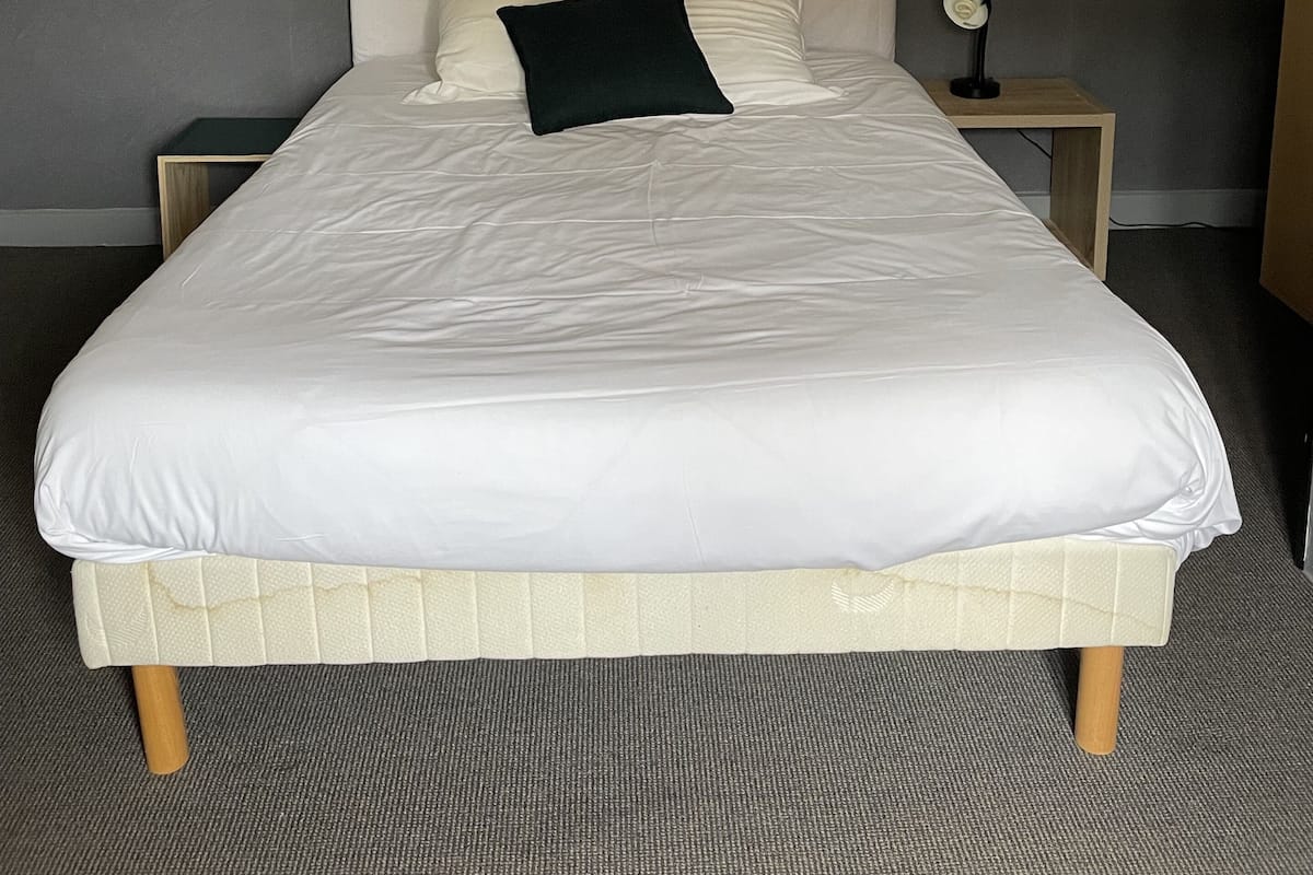 A neatly made bed with white linens and a black pillow, a bedside table with a lamp, and a woven light fixture above.