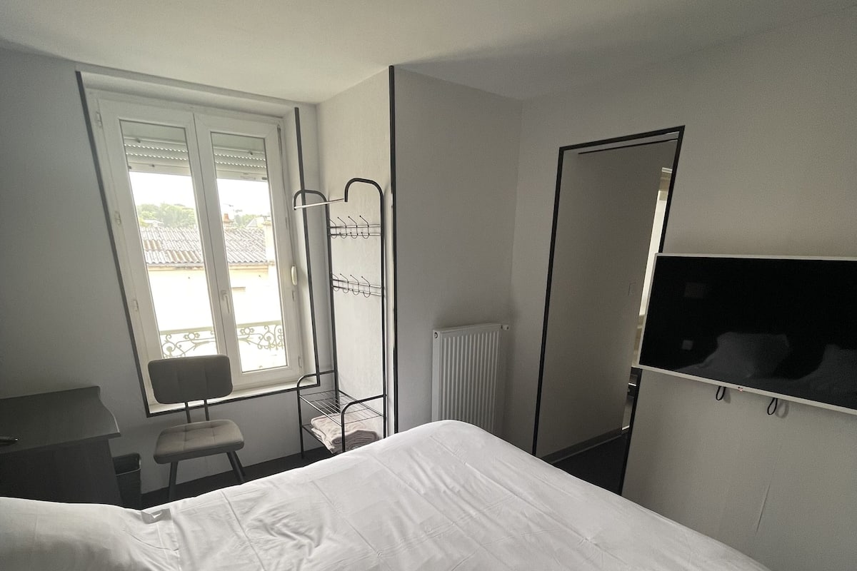 A hotel room with a large bed, a desk, a chair, a radiator, and a window with blinds.