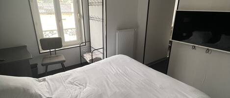 A hotel room with a large bed, a desk, a chair, a radiator, and a window with blinds.