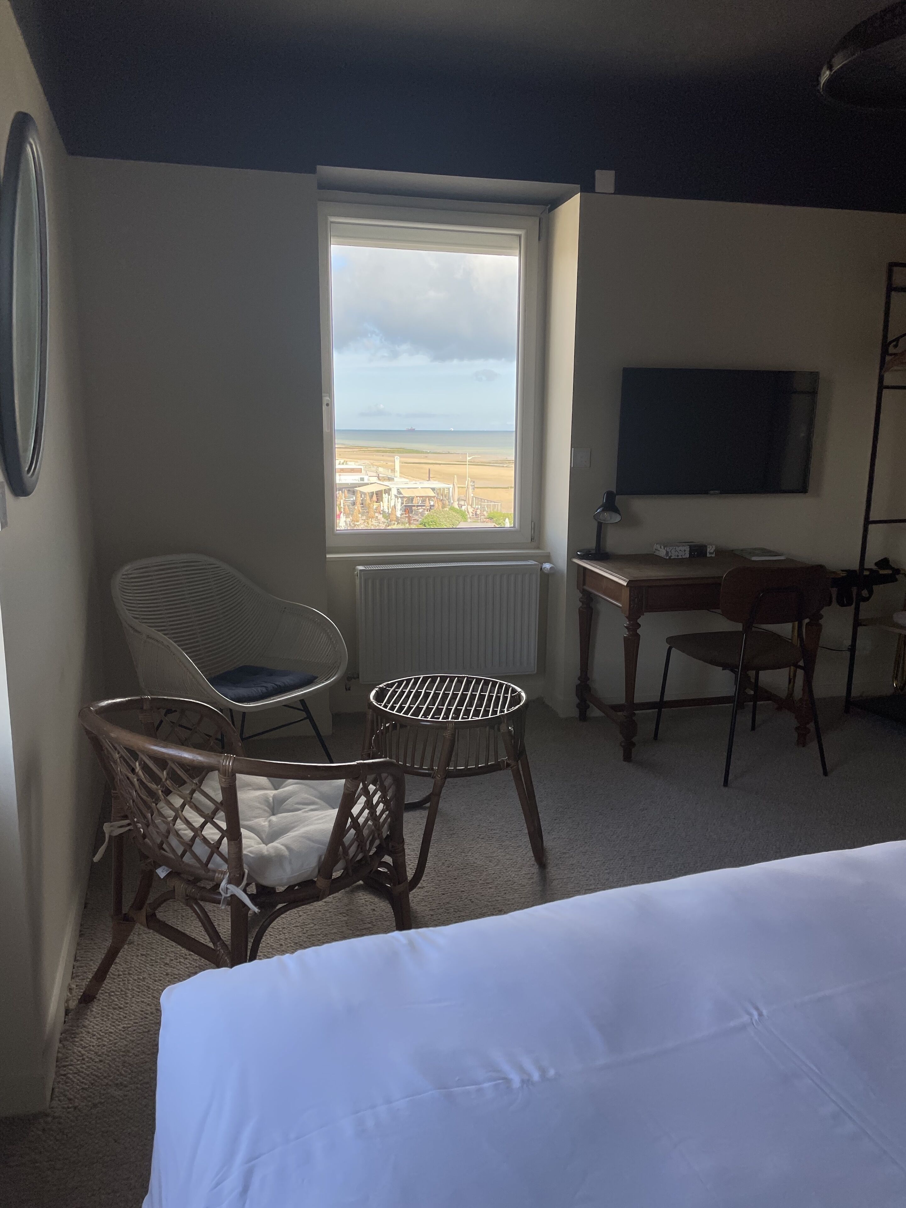 Une chambre d’hôtel avec un lit, un bureau, deux chaises, une grande fenêtre donnant sur la plage et un téléviseur fixé au mur.