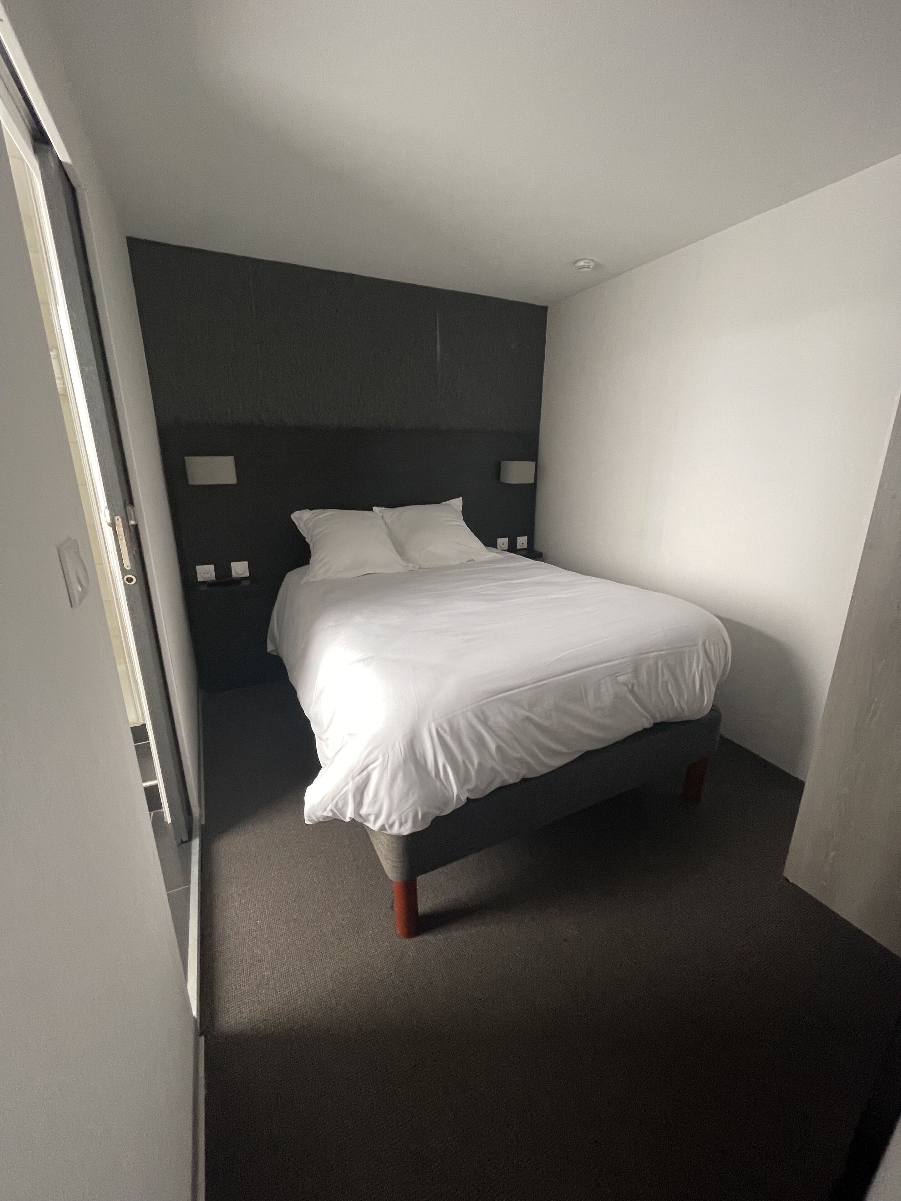 A small, modern hotel room with a single bed, a dark headboard, and a bedside table with a lamp.