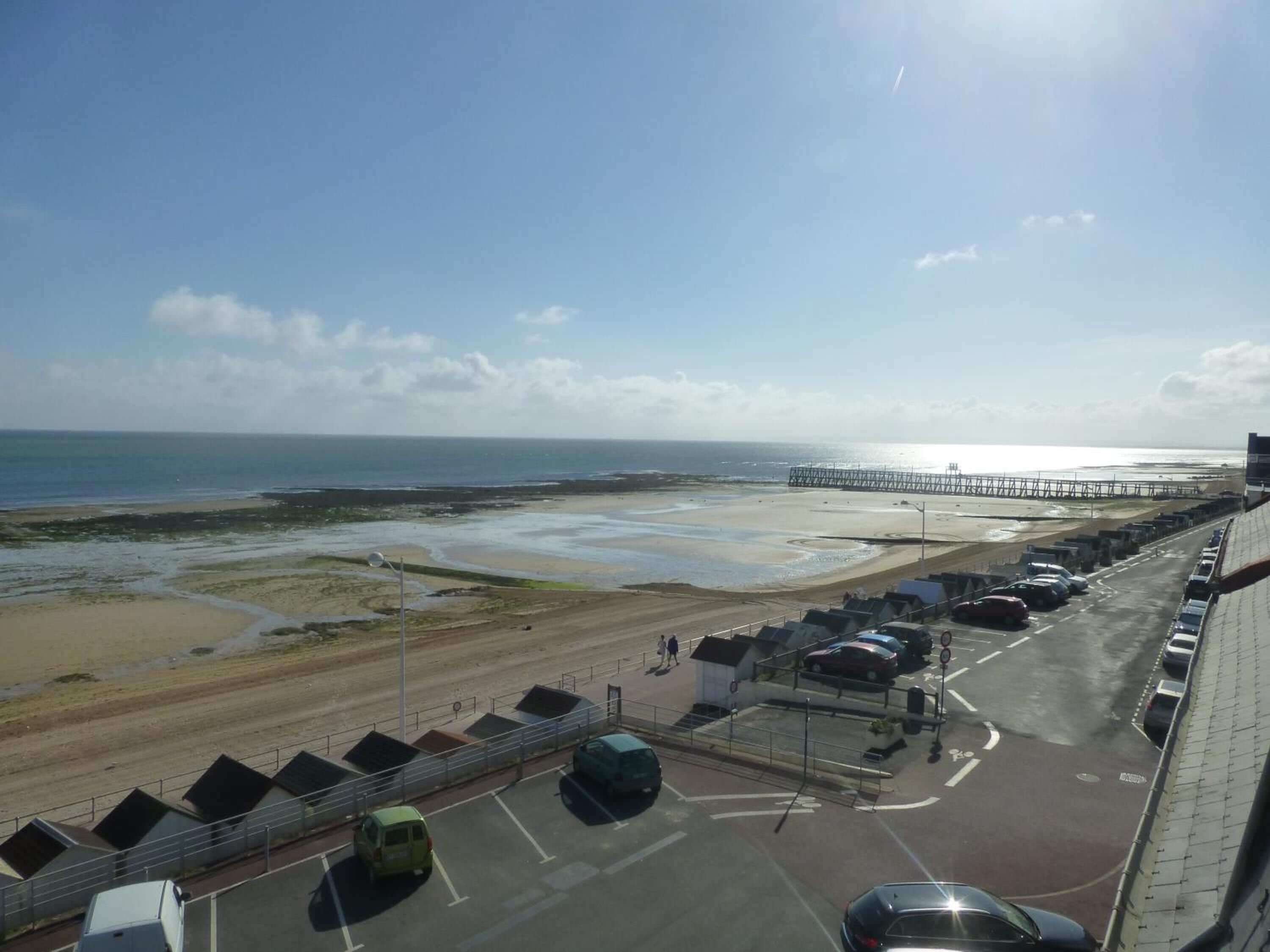 A coastal view with a beach, a pier, and a parking area with cars.