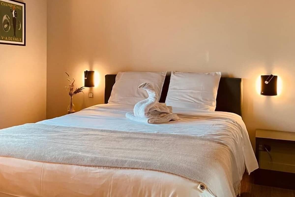 A neatly made bed with a towel, a framed poster on the wall, and wall-mounted lights.