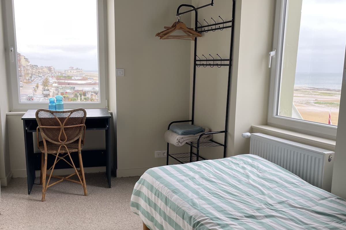 A bedroom with a bed, a desk with a chair, a radiator, and a window with a view of the beach.