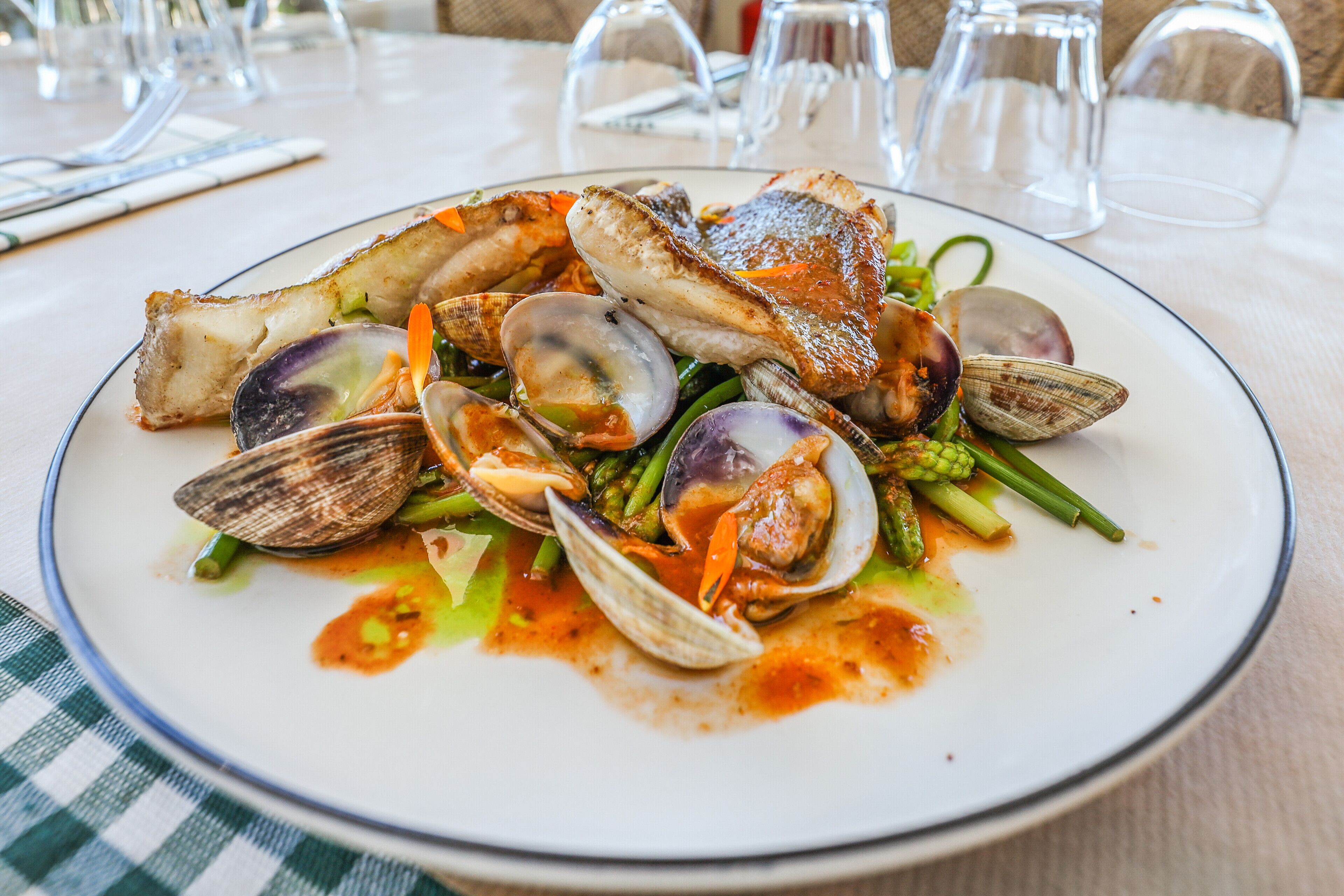 Un plat de fruits de mer, composé de palourdes, d’asperges et d’un morceau de poisson, est servi sur une table recouverte d’une nappe à carreaux.