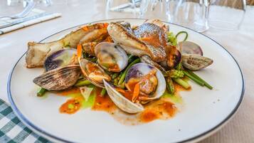 Un plat de fruits de mer, composé de palourdes, d’asperges et d’un morceau de poisson, est servi sur une table recouverte d’une nappe à carreaux.