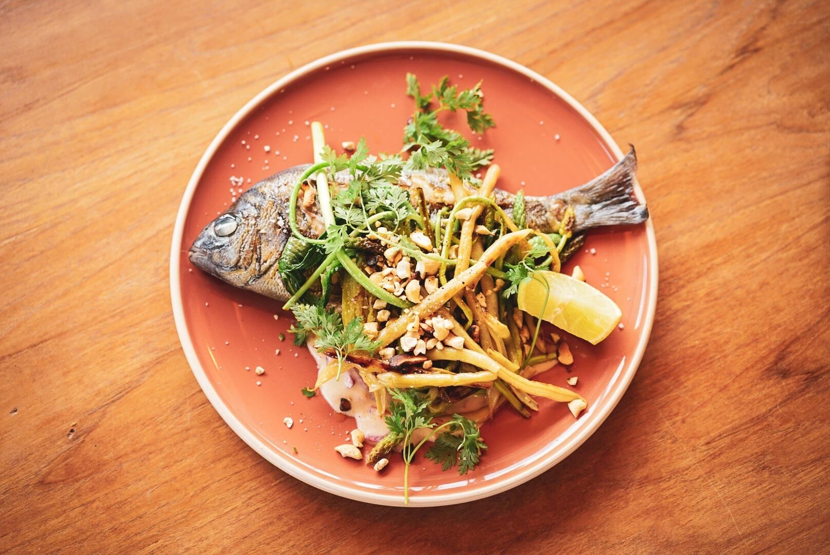 A plate of food with a fish, vegetables, and nuts on a wooden table.