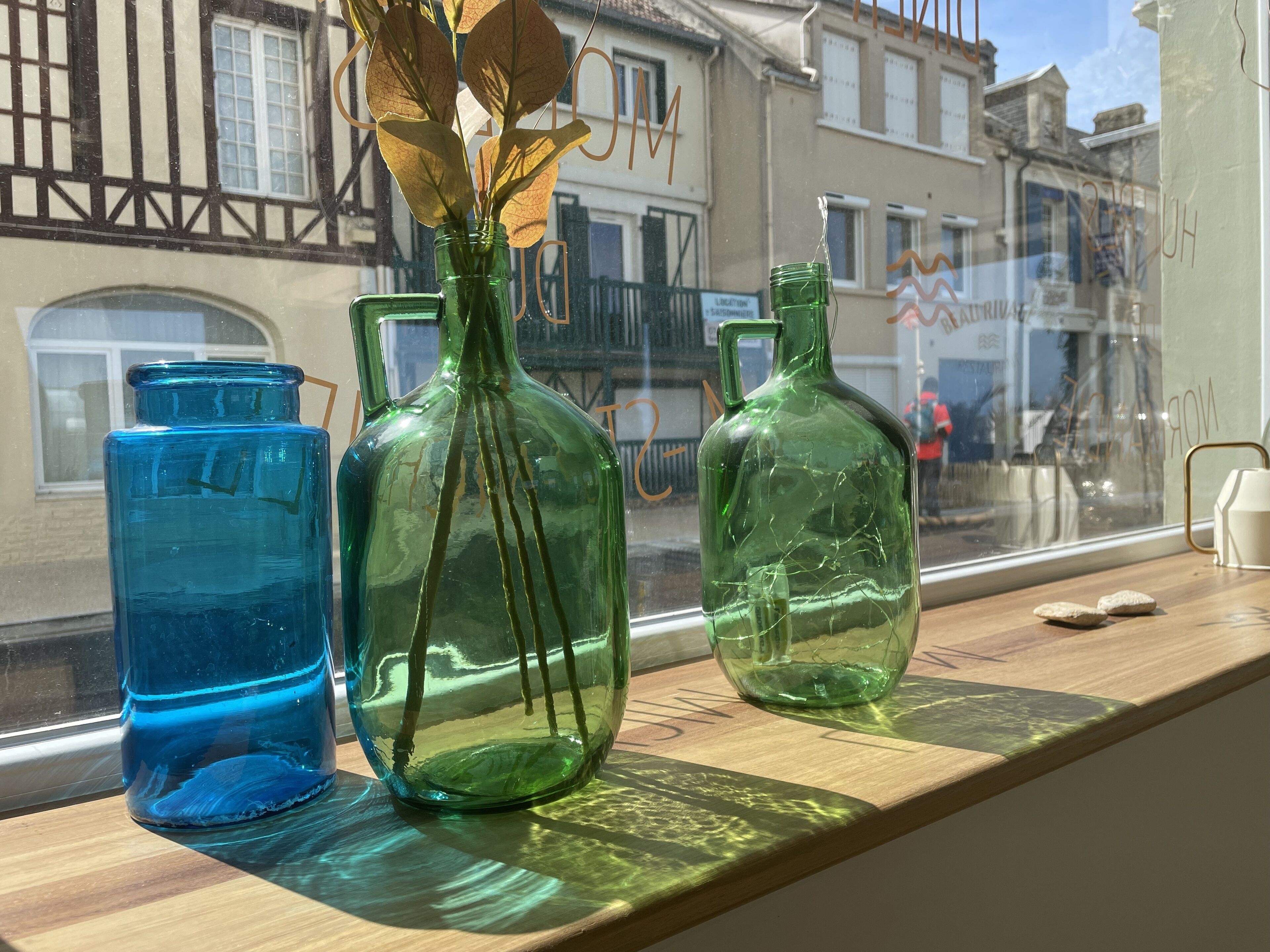 Three glass vases with dried leaves on a wooden shelf by a window.