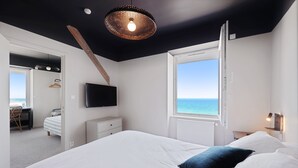 A bedroom with a large bed, a TV mounted on the wall, a window with a view of the sea, and a ceiling with a unique light fixture. - Le Beau Rivage (Luc-sur-Mer)