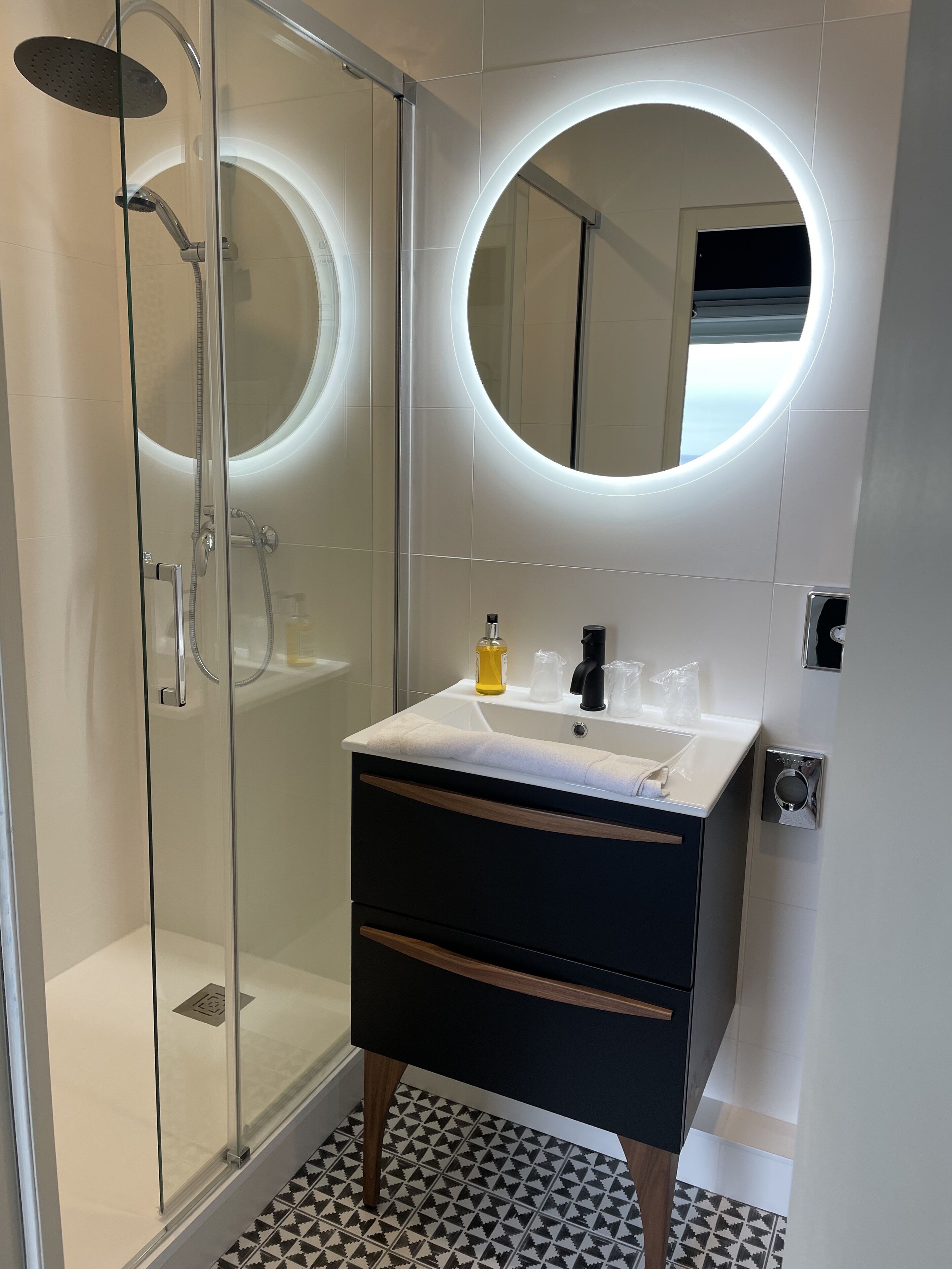 A modern bathroom with a glass-enclosed shower, a round mirror with a light, a white sink, and a black vanity with drawers.