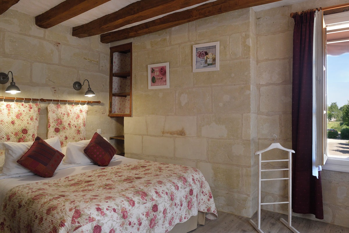 A bedroom with a floral bedspread, a wooden headboard, a hanging light fixture, and a window with a view of greenery.