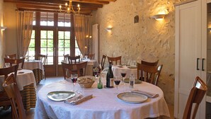 A dining area with round tables, wooden chairs, and a stone wall. - Auberge Le Colombien - Hôtel Restaurant (Villandry)