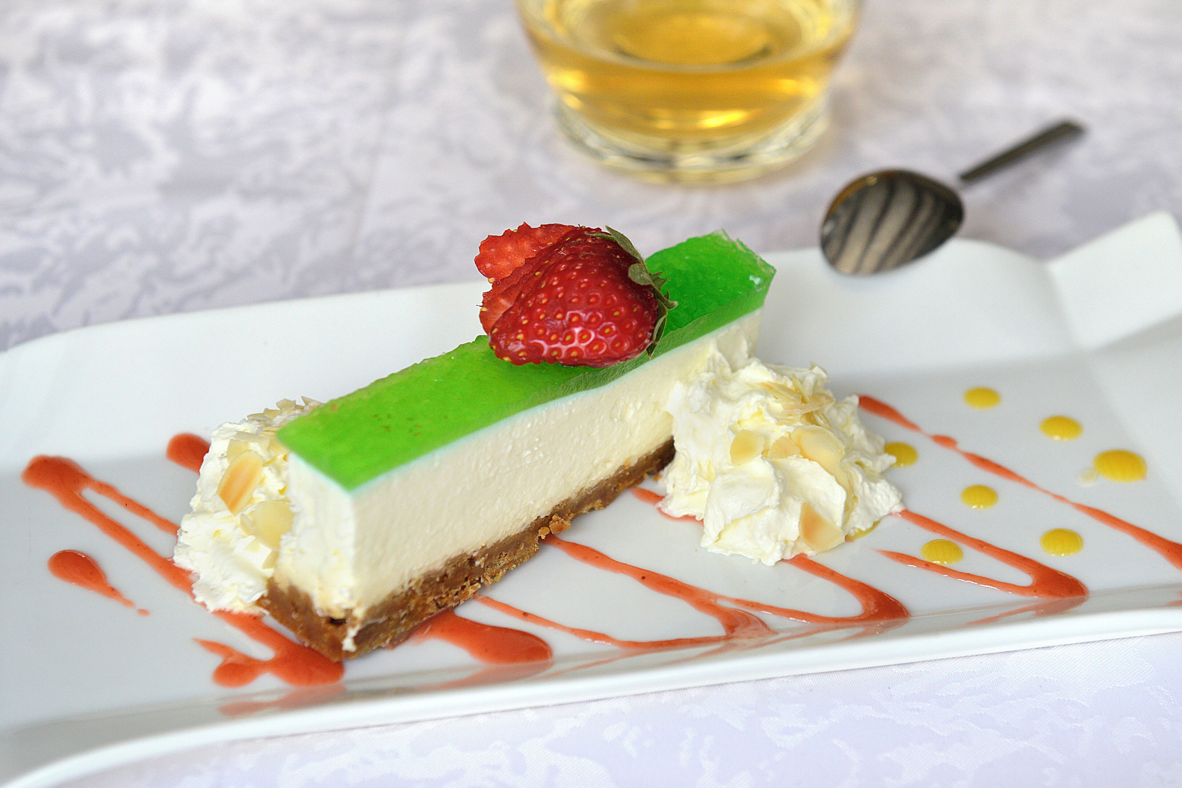 Una rebanada de pastel de queso con fresa encima, rociada con salsa roja y amarilla, sobre un plato blanco.