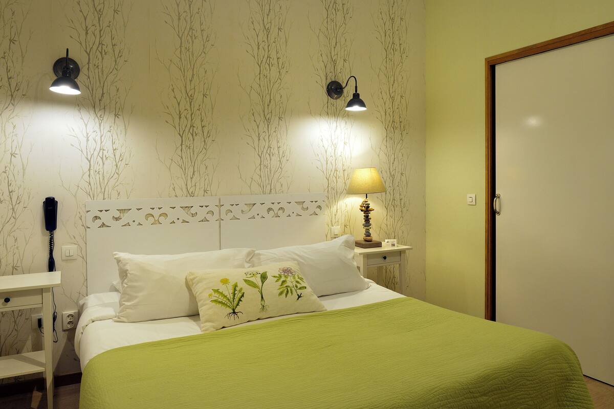 A neatly made bed with a green bedspread, two bedside lamps, and a wall with a nature-themed wallpaper.